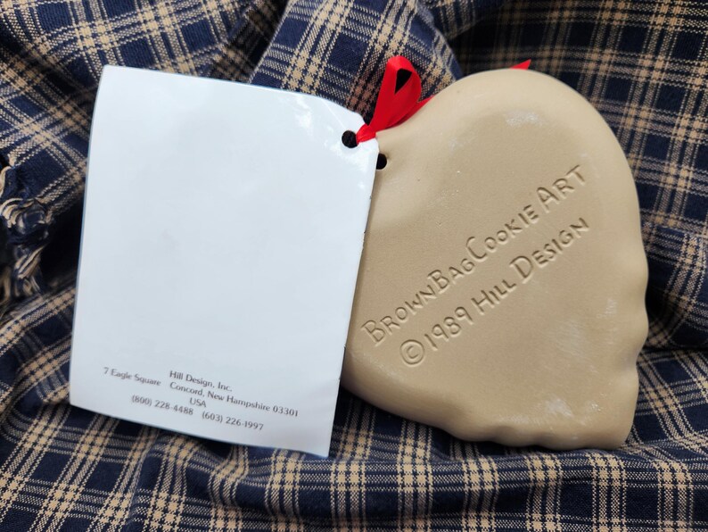May include: A tan heart-shaped ceramic cookie mold with a red ribbon, featuring the embossed text BrownBagCookieArt 1989 Hill Design. Attached is a white paper tag with the address 7 Eagle Square, Concord, New Hampshire 03301 and phone numbers.