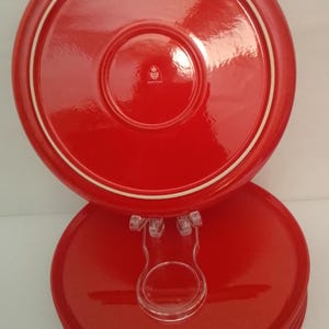 May include: A stack of round, glossy red ceramic plates with a white rim accent. The top plate features a small, centered white emblem with a crown and shield design. The plates are held in place by a clear plastic display stand for visibility.