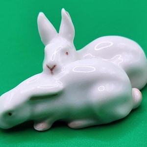 May include: A white porcelain figurine featuring two rabbits in a resting position. One rabbit is lying down while the other sits upright behind it. The rabbits have smooth, glossy surfaces with subtle pink accents on their noses and small tails.
