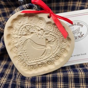 May include: A heart-shaped beige ceramic cookie mold featuring an embossed design of two birds and floral patterns. A bright red ribbon is tied through a hole at the top. Beside it is a white booklet labeled Crown Bag Cookie Art.