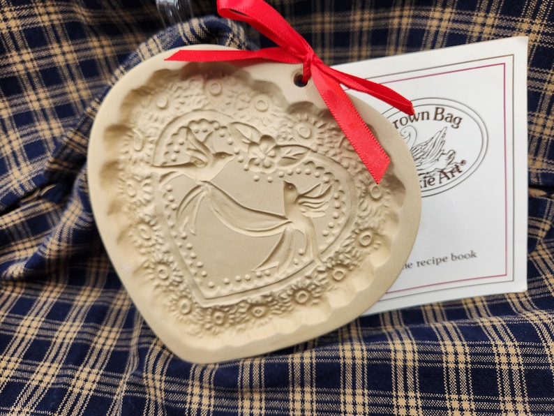 May include: A heart-shaped beige ceramic cookie mold featuring an embossed design of two birds and floral patterns. A bright red ribbon is tied through a hole at the top. Beside it is a white booklet labeled Crown Bag Cookie Art.