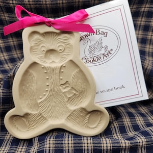 May include: A beige ceramic bear-shaped cookie mold featuring a detailed embossed teddy bear design, accented with a bright pink ribbon tied at the top. Beside it is a white booklet with the text Brown Bag Cookie Art and a swan illustration.