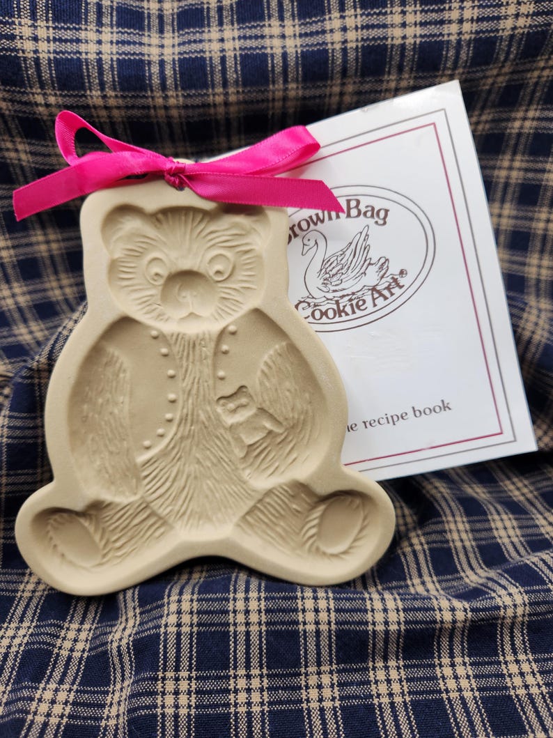 May include: A beige ceramic bear-shaped cookie mold featuring a detailed embossed teddy bear design, accented with a bright pink ribbon tied at the top. Beside it is a white booklet with the text Brown Bag Cookie Art and a swan illustration.