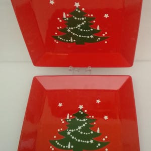 May include: Two square red ceramic plates featuring a centered green Christmas tree graphic. The tree is decorated with white dots and small white stars scattered across the red background. The plates have a smooth, glossy finish and raised edges.