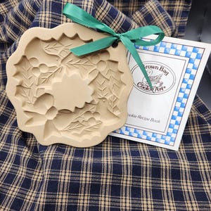 May include: A beige ceramic cookie mold with a holly leaf and berry design, featuring a green ribbon tied through a small hole at the top. Beside it is a white booklet with blue checkered borders and the text Brown Bag Cookie Art Cookie Recipe Book.
