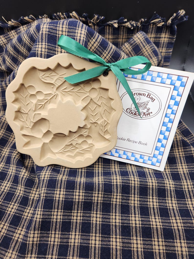 May include: A beige ceramic cookie mold with a holly leaf and berry design, featuring a green ribbon tied through a small hole at the top. Beside it is a white booklet with blue checkered borders and the text Brown Bag Cookie Art Cookie Recipe Book.