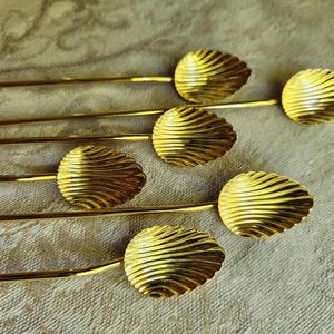 May include: Six gold-colored serving spoons with a leaf-shaped design on the spoon head. The spoons have a long, slender handle and a textured, ridged pattern on the spoon head. The spoons are arranged on a light-colored, textured surface.