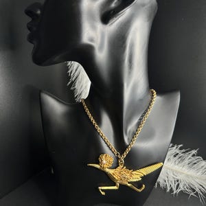 May include: A gold-colored necklace featuring a roadrunner pendant. The pendant is detailed with textured feathers and a small, round embellishment. The necklace is displayed on a black mannequin, with a chain-link style necklace.