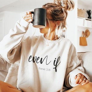 Può includere: Una donna indossa una felpa beige chiaro con il testo "even if" e "Daniel 3:18" in lettere nere. È seduta su un divano grigio chiaro con le gambe incrociate e tiene una tazza nera.