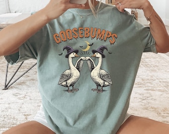 May include: A pale green t-shirt with the word "GOOSEBUMPS" in orange lettering. The shirt features a graphic of two geese wearing witch hats, with a crescent moon and bats. The t-shirt has a relaxed fit.