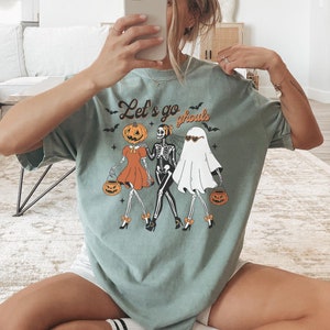 May include: Sage green t-shirt with the text "Let's go ghouls" and a graphic of a pumpkin, skeleton, and ghost holding jack-o'-lanterns. The shirt has a relaxed fit and is perfect for Halloween.