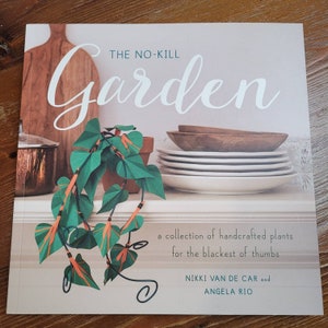 May include: The cover of a book titled "The No-Kill Garden: A collection of handcrafted plants for the blackest of thumbs" by Nikki Van De Car and Angela Rio. The cover features a green plant with orange accents against a light background.