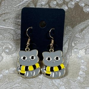 May include: A pair of gold cat earrings with a gray cat face and a yellow and black striped scarf. The earrings are hanging from a black card.