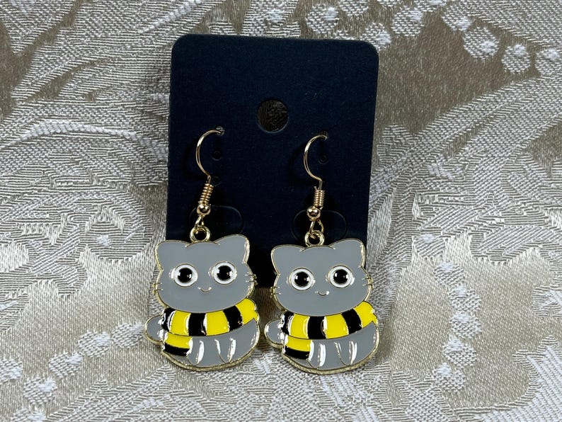 May include: A pair of gold cat earrings with a gray cat face and a yellow and black striped scarf. The earrings are hanging from a black card.