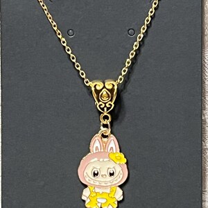 May include: Gold-colored chain necklace with a pendant featuring a cartoon bunny character. The bunny is wearing a pink hood and yellow overalls. The pendant is approximately 1 inch in height.