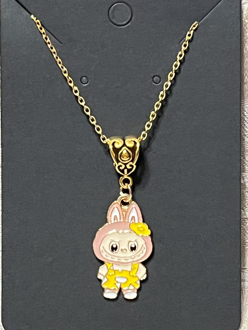 May include: Gold-colored chain necklace with a pendant featuring a cartoon bunny character. The bunny is wearing a pink hood and yellow overalls. The pendant is approximately 1 inch in height.