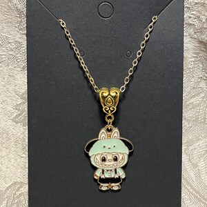 May include: A gold-colored chain necklace with a pendant featuring a cartoon character in a light green hooded outfit. The pendant has black and white details, and the necklace is displayed on a black card.