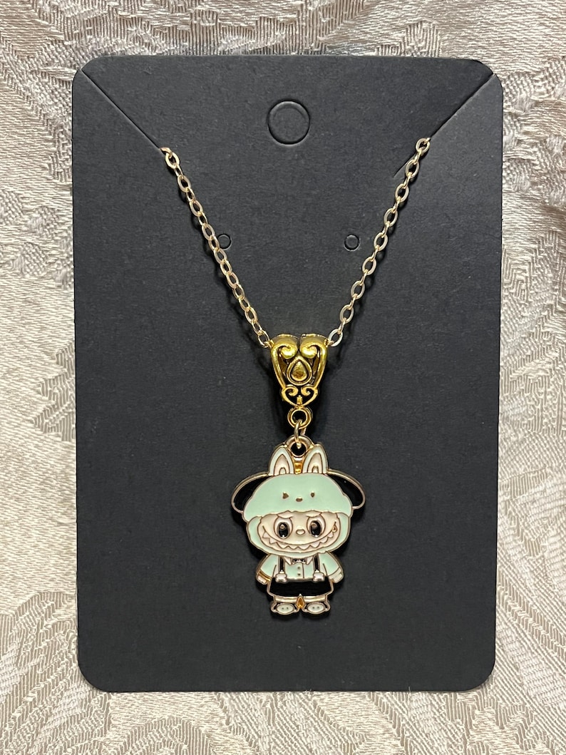 May include: A gold-colored chain necklace with a pendant featuring a cartoon character in a light green hooded outfit. The pendant has black and white details, and the necklace is displayed on a black card.