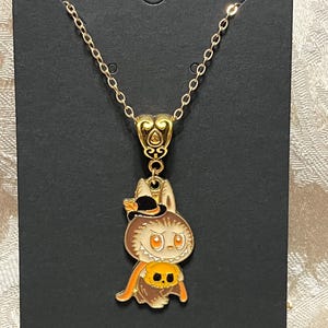 May include: Gold-toned necklace with a Halloween-themed pendant. The pendant features a cartoon bunny wearing a witch hat and holding a pumpkin. The bunny is brown, orange, and white, with black accents. The necklace is displayed on a black card.