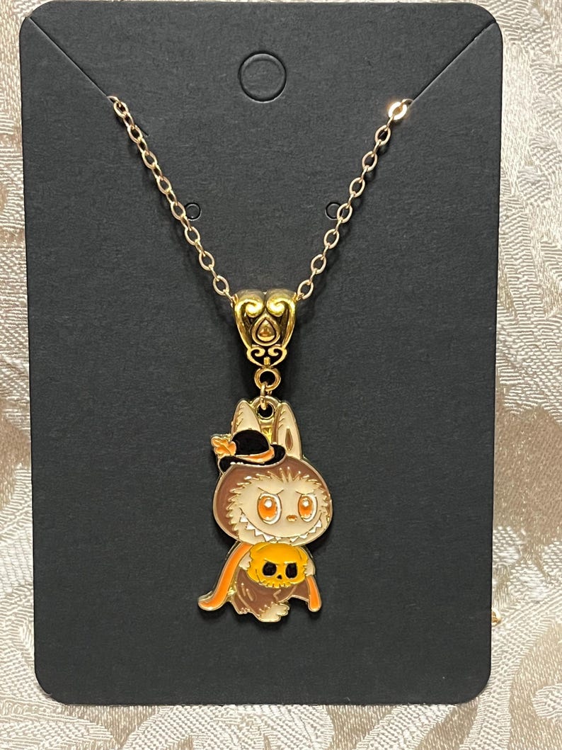 May include: Gold-toned necklace with a Halloween-themed pendant. The pendant features a cartoon bunny wearing a witch hat and holding a pumpkin. The bunny is brown, orange, and white, with black accents. The necklace is displayed on a black card.