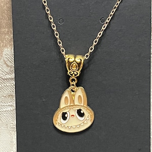 May include: Gold-toned necklace featuring a pendant with a cartoon bunny design. The bunny has a cream-colored face, large black eyes, and a light brown hat. The necklace is displayed on a black card.