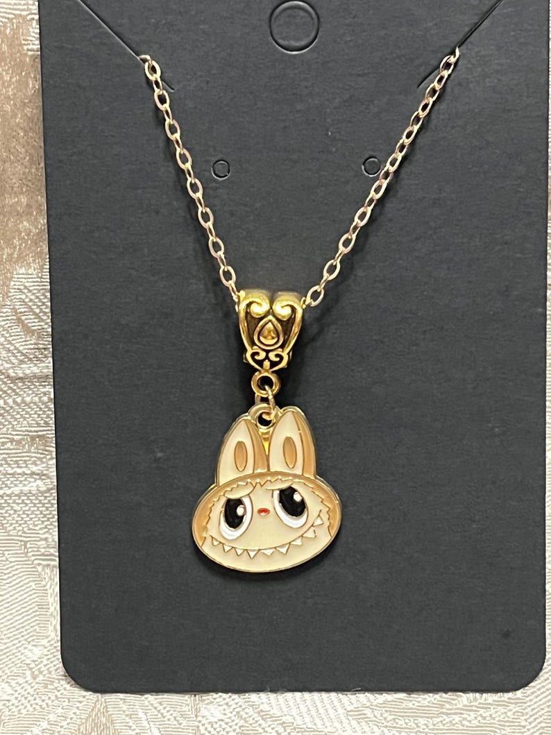 May include: Gold-toned necklace featuring a pendant with a cartoon bunny design. The bunny has a cream-colored face, large black eyes, and a light brown hat. The necklace is displayed on a black card.