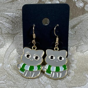 May include: A pair of dangle earrings featuring a gray cat wearing a green and white striped scarf. The cat has black eyes and a gold outline.
