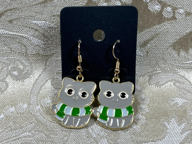 May include: A pair of dangle earrings featuring a gray cat wearing a green and white striped scarf. The cat has black eyes and a gold outline.