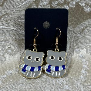 May include: A pair of gray cat-shaped earrings with blue and white striped scarves. The earrings are made of metal and have gold-colored hooks.