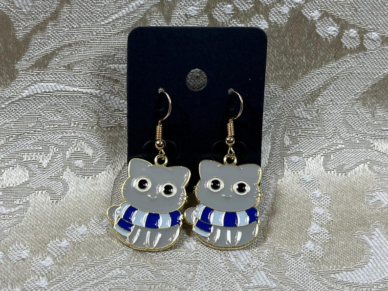 May include: A pair of gray cat-shaped earrings with blue and white striped scarves. The earrings are made of metal and have gold-colored hooks.