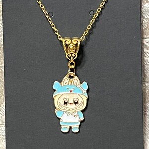 May include: Gold-toned necklace featuring a pendant of a cartoon character in a blue outfit with a matching hat. The pendant is detailed with white and light blue enamel. The necklace has a delicate chain and a decorative bail.