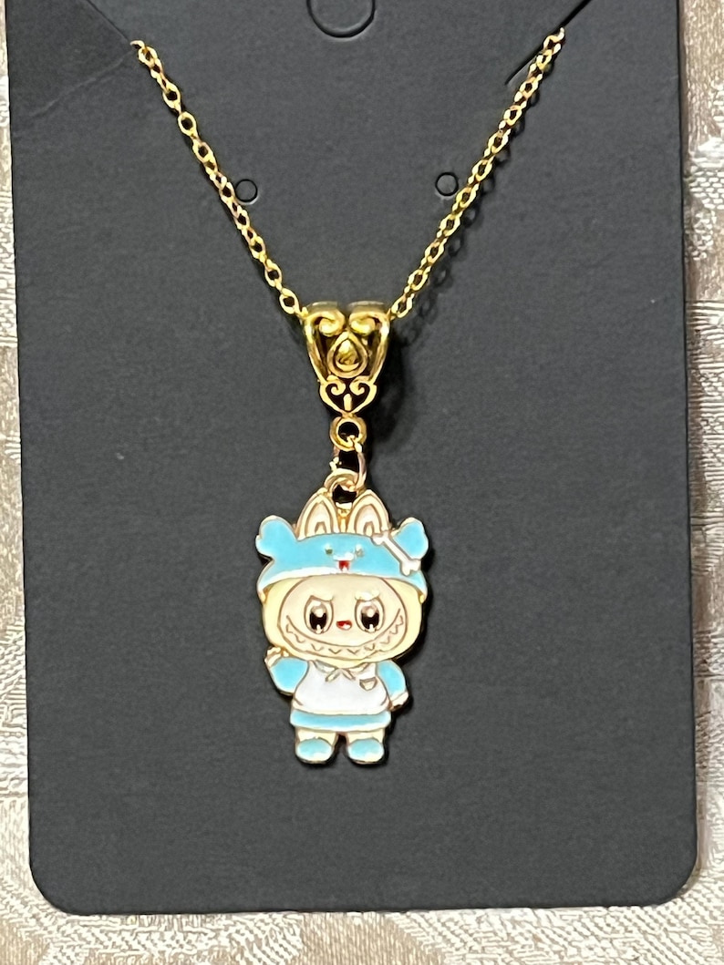 May include: Gold-toned necklace featuring a pendant of a cartoon character in a blue outfit with a matching hat. The pendant is detailed with white and light blue enamel. The necklace has a delicate chain and a decorative bail.