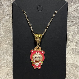 May include: A gold-colored chain necklace with a pendant featuring a cartoon bear in a red hooded outfit. The pendant has a heart-shaped bail and the bear is holding a strawberry. The necklace is displayed on a black card.
