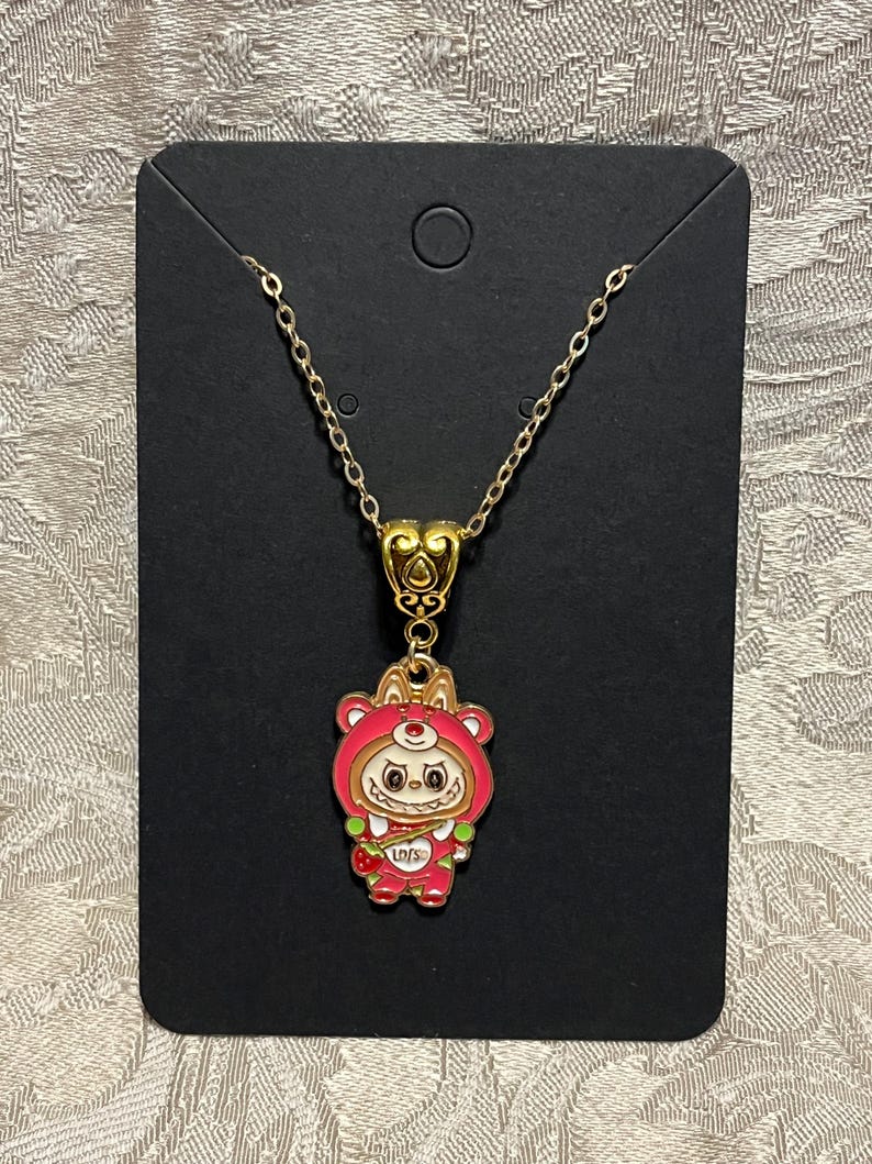 May include: A gold-colored chain necklace with a pendant featuring a cartoon bear in a red hooded outfit. The pendant has a heart-shaped bail and the bear is holding a strawberry. The necklace is displayed on a black card.