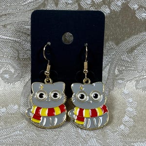 May include: A pair of dangle earrings featuring a gray cat wearing a red and yellow scarf. The cat has large, round eyes and a small, gold star on its forehead.