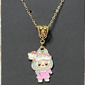 May include: Gold-colored necklace featuring a pendant with a cartoon character in a pink dress and blue accents, holding a wand. A small Hello Kitty figure is on the character's shoulder. The necklace is on a black background.