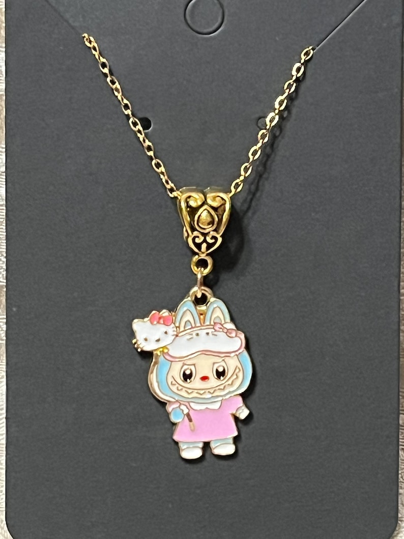May include: Gold-colored necklace featuring a pendant with a cartoon character in a pink dress and blue accents, holding a wand. A small Hello Kitty figure is on the character's shoulder. The necklace is on a black background.