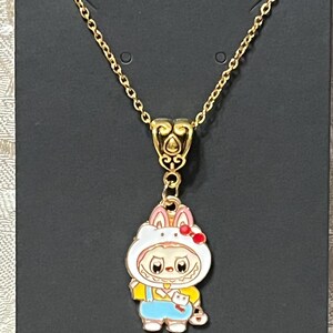 May include: Gold-colored necklace with a pendant featuring a cartoon character in a bunny costume. The pendant has a white bunny hood with pink ears and a red bow. The character wears a yellow shirt and blue overalls.