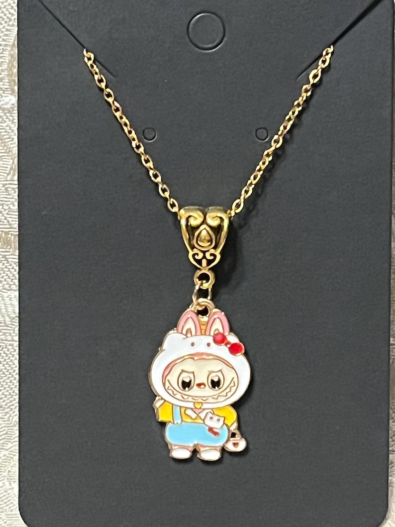 May include: Gold-colored necklace with a pendant featuring a cartoon character in a bunny costume. The pendant has a white bunny hood with pink ears and a red bow. The character wears a yellow shirt and blue overalls.