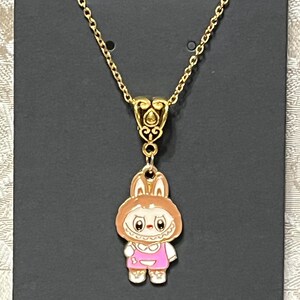 May include: Gold-toned necklace featuring a pendant of a cartoon bunny wearing a pink outfit. The bunny pendant has a white face, brown ears, and is suspended from a delicate chain. The necklace is displayed on a black card.