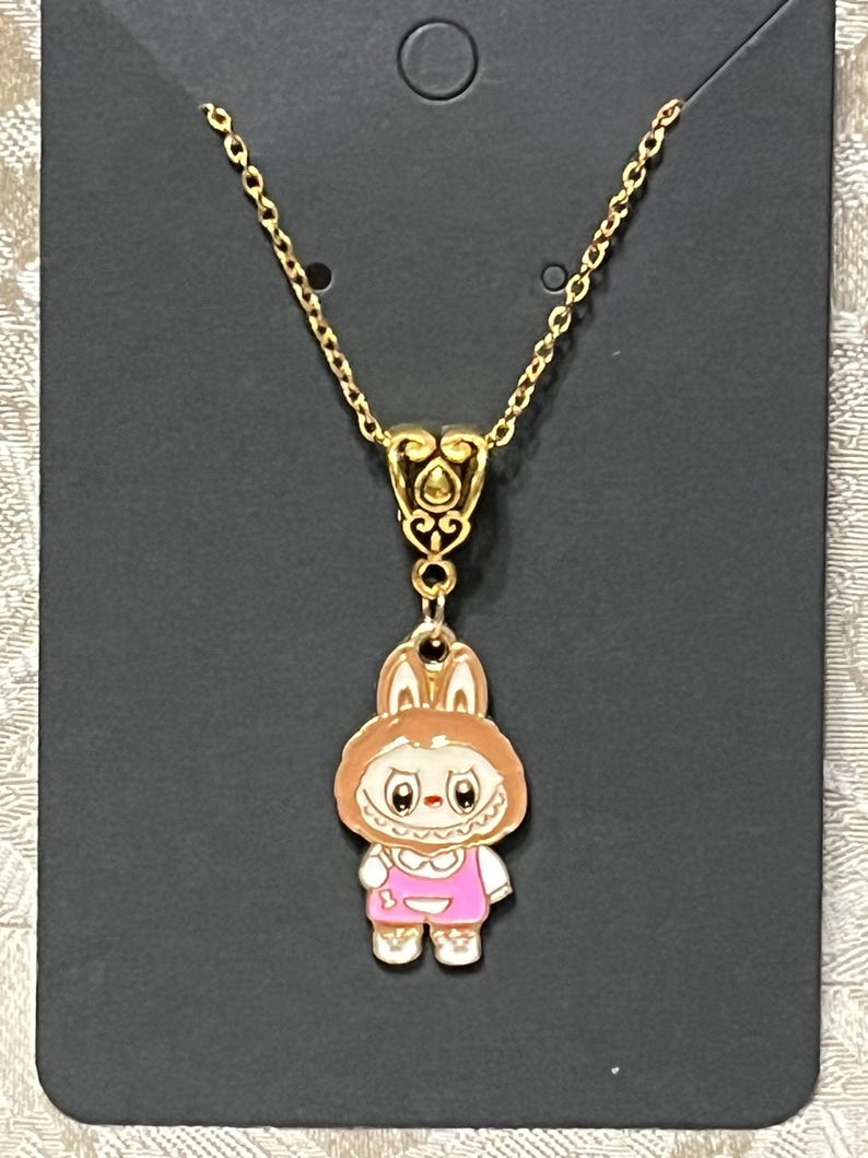 May include: Gold-toned necklace featuring a pendant of a cartoon bunny wearing a pink outfit. The bunny pendant has a white face, brown ears, and is suspended from a delicate chain. The necklace is displayed on a black card.