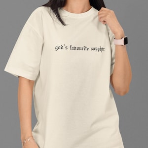 May include: Cream-coloured t-shirt with the text "god's favourite sapphic" in black gothic font. The shirt is a relaxed fit. The person is wearing grey cargo trousers and a pink watch.