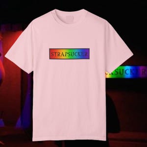 May include: A light pink t-shirt with the word "STRAPSUCKER" in a rainbow-colored rectangular design across the chest. The text is outlined in black. The shirt has short sleeves and a crew neck.