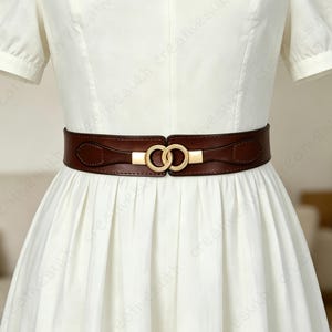 May include: A dark brown waist belt featuring a gold-toned interlocking double ring buckle. The belt is worn over a plain white dress with short sleeves and a pleated skirt. The design includes smooth leather-like material with stitched details.