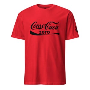 May include: A red t-shirt featuring the words "Come Caca zero" in black, styled after a well-known brand's logo. The tee has a crew neck and short sleeves. A small logo is on the right sleeve.