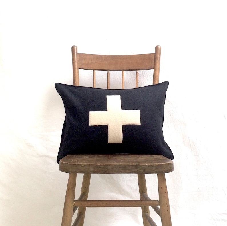 Puede incluir: Una almohada de tiro negra y blanca con un dise&ntilde;o de cruz blanca. La almohada est&aacute; sentada en una silla de madera.