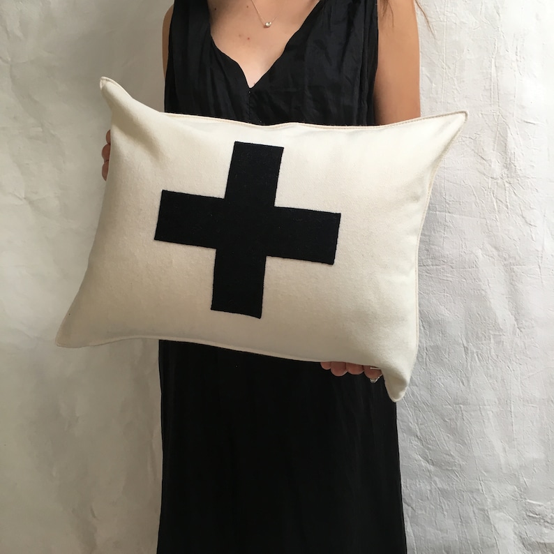 Puede incluir: Una almohada blanca con un gran signo m&aacute;s negro en el centro.