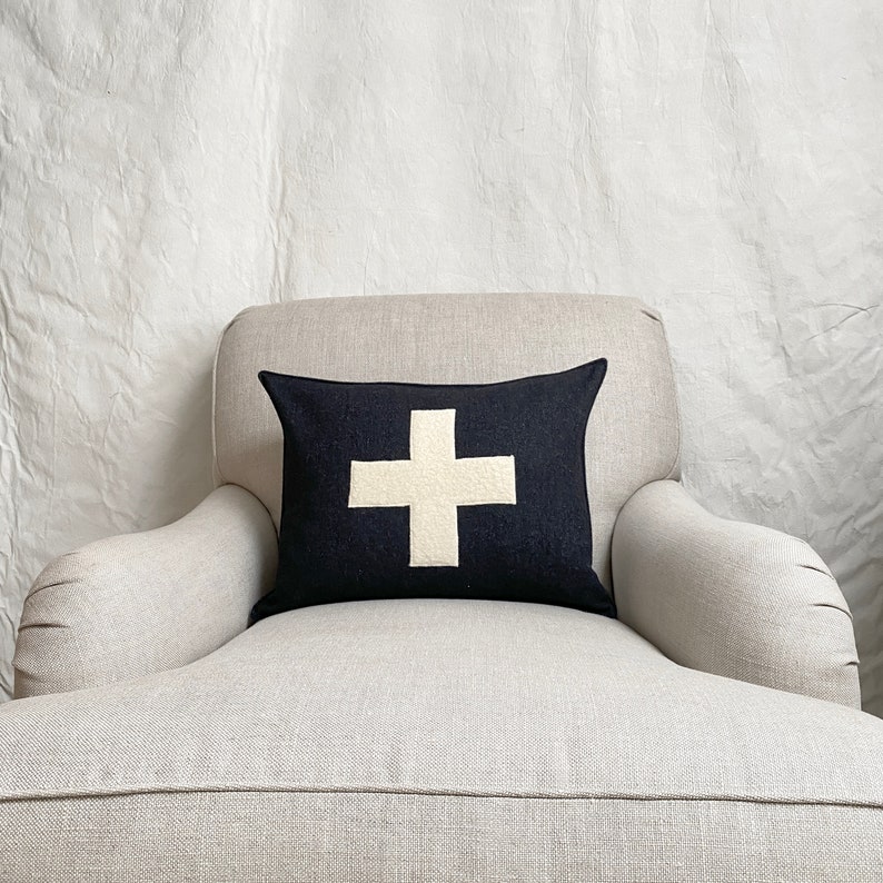 Puede incluir: Un coj&iacute;n negro con una cruz blanca sobre un sill&oacute;n gris claro. El sill&oacute;n est&aacute; frente a una pared blanca.