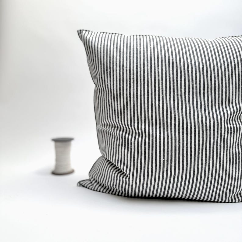 Puede incluir: Un coj&iacute;n cuadrado con un patr&oacute;n de rayas blancas y negras. El coj&iacute;n es de tela y tiene un aspecto suave y afelpado. Una bobina de hilo est&aacute; en el fondo. El coj&iacute;n es un elemento decorativo para una cama o un sof&aacute;.