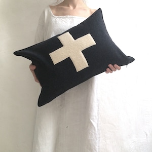 Puede incluir: Una almohada negra con un dise&ntilde;o de cruz de fieltro blanco. La almohada est&aacute; siendo sostenida por una persona que lleva un vestido blanco.